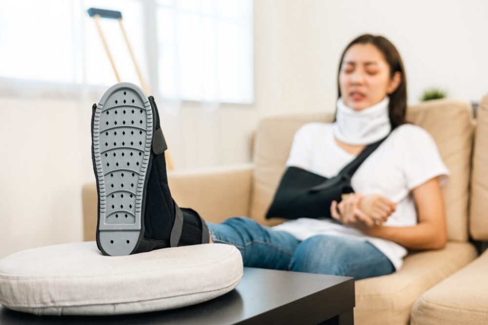woman with neck brace, arm sling and foot in a cast sitting on the couch