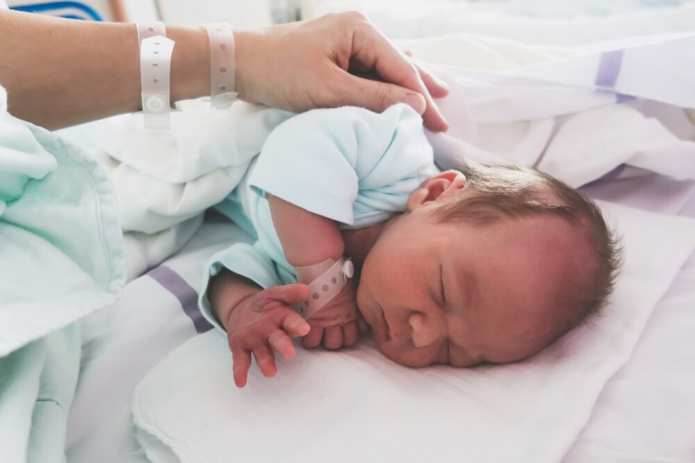 An infant laying on it's side in a hospital with it's parents hand resting on it's back.