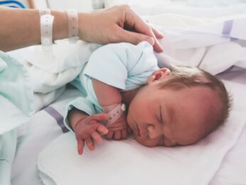 An infant laying on it's side in a hospital with it's parents hand resting on it's back.