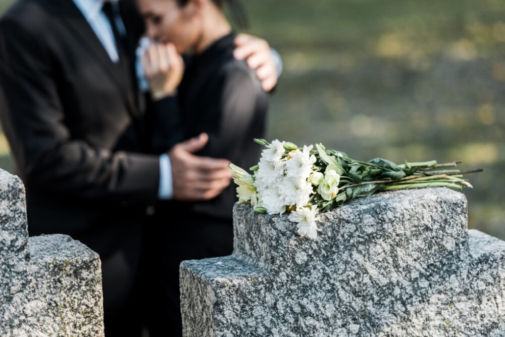 Two people dressed in black and hugging in front of a headstone in a cemetary.