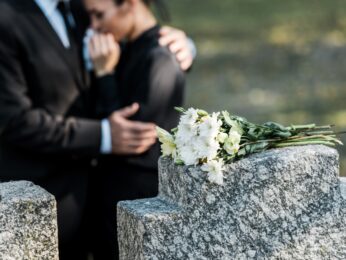 Two people dressed in black and hugging in front of a headstone in a cemetary.